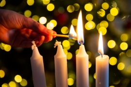 Eine Hand zündet die zweite von vier weißen Kerzen an. Eine Kerze brennt bereits.