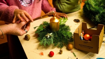 Auf einem Tisch steht ein Gesteck mit Tujazweigen und einer goldenen Kerze. Daneben kleine Weihnachtskugeln. Eine Kinderhand und eine Erwachsenenhand halten einen Stock.
